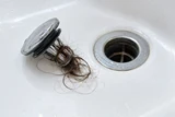 A removed bathtub drain stopper with tangled hair beside the empty drain opening in a white tub