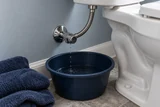 A small leak at a toilet supply hose captured in a shallow bucket below a chrome angle stop; dry towels are nearby