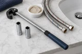 A basin wrench and slim deep sockets laid beside braided supply hoses and plumbers putty on a pale bathroom work surface