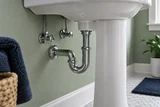A white ceramic pedestal sink viewed low from the side, showing its chrome P-trap and shutoff valves against a sage bathroom wall