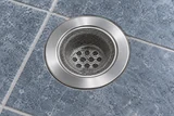 A reusable stainless hair-catching screen above a clean shower drain, with the fitted screen and tiled floor as the main subjects