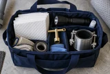An emergency plumbing kit with shutoff key, absorbent pads, torch and pipe repair clamp neatly arranged in a navy tote