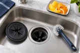 A garbage disposal splash guard removed from the sink opening beside a long-handled cleaning brush, citrus peel on a small tray