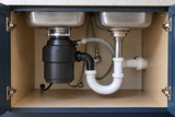 A garbage disposal installed beneath a double-bowl kitchen sink, with drain elbow and dishwasher connection visible in a tidy open cabinet