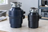 Two uninstalled garbage disposals of different sizes standing on a pale workshop surface, one showing its mounting ring