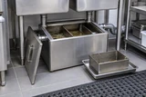 An opened stainless commercial grease interceptor below a prep sink, its removable basket resting on a tray