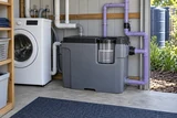 A compact greywater collection tank with filtration basket and lilac reuse pipes beside laundry plumbing in a utility shed