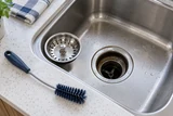 A stainless kitchen basket strainer lifted out to expose the drain opening, with a short flexible cleaning brush on the counter