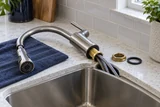 A pull-down kitchen tap being replaced: faucet base and mounting nut beside the sink cutout, flexible hoses visible