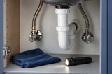 Two kitchen sink supply angle stops and supply hoses being inspected, a dry cloth and small flashlight resting below