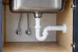 The underside of a deep stainless kitchen sink with a white PVC P-trap and two neat supply lines in an open cabinet