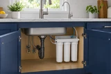 A three-canister under-sink drinking-water filtration unit with small tubing and a dedicated tap above in a bright kitchen