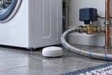 A small smart water leak sensor on the floor beside a washing-machine supply hose, with a connected automatic shutoff valve in the background