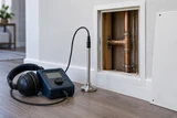 An acoustic leak detector and listening probe positioned beside a concealed copper pipe access panel in a dry household wall