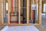 Open residential timber wall framing with neat red and blue PEX branches, secured supports and a plumbing plan resting below
