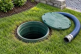A septic tank inspection riser open in a lawn, with an inspection hose resting beside the green lid