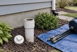 A sewer cleanout outside a house with its cap removed, drain rods and containment tarp beside it