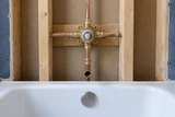 An exposed brass shower mixing valve and copper risers in an unfinished bathtub wall, with the clean white tub rim below