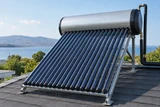An evacuated-tube solar water heater mounted on a low pitched roof, repeating dark glass tubes and small storage tank visible beneath clear sky