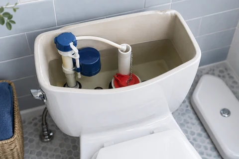 An open white toilet cistern showing the fill valve, float and red flapper, with the lid resting beside it in a pale tiled bathroom