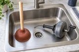 A manual cup plunger standing in a kitchen sink next to a hand-operated drain auger