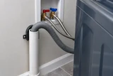 A laundry washer standpipe and drain hose with red and blue supply connections visible behind a washing machine