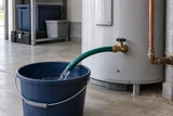 A garden hose attached to a water heater drain valve for flushing, directed toward a bucket in a clean garage