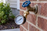 A water pressure gauge screwed onto an outdoor hose bib against brick, the circular face and metal fitting clearly visible