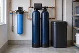 A twin-cylinder water softener and brine tank beside a whole-house filter in a neat utility room